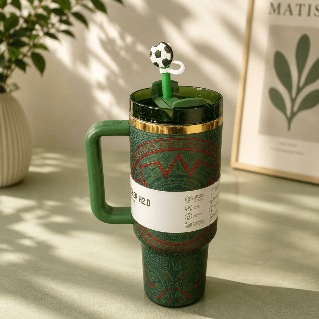 Green travel mug with soccer-themed lid on a light surface with a plant and framed picture in the background.