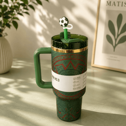 Green travel mug with soccer-themed lid on a light surface with a plant and framed picture in the background.