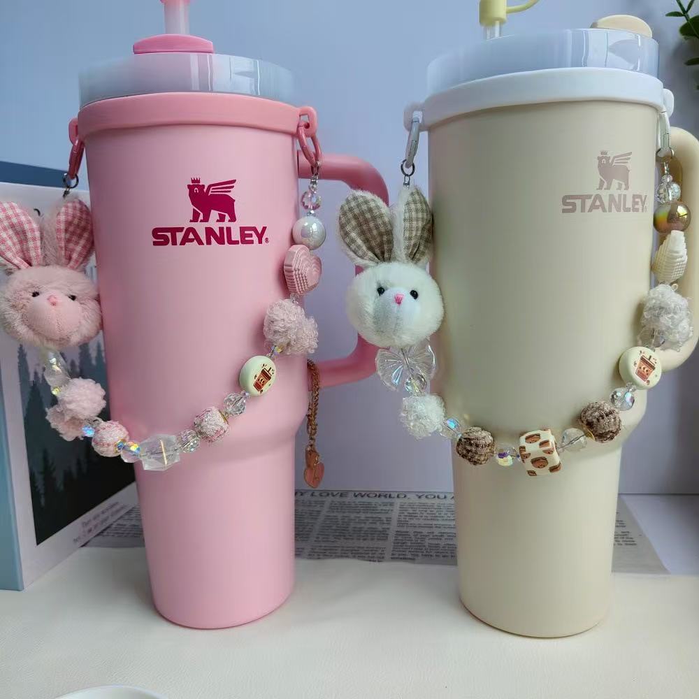 Pink and beige Stanley tumblers with decorative charms on a light blue background