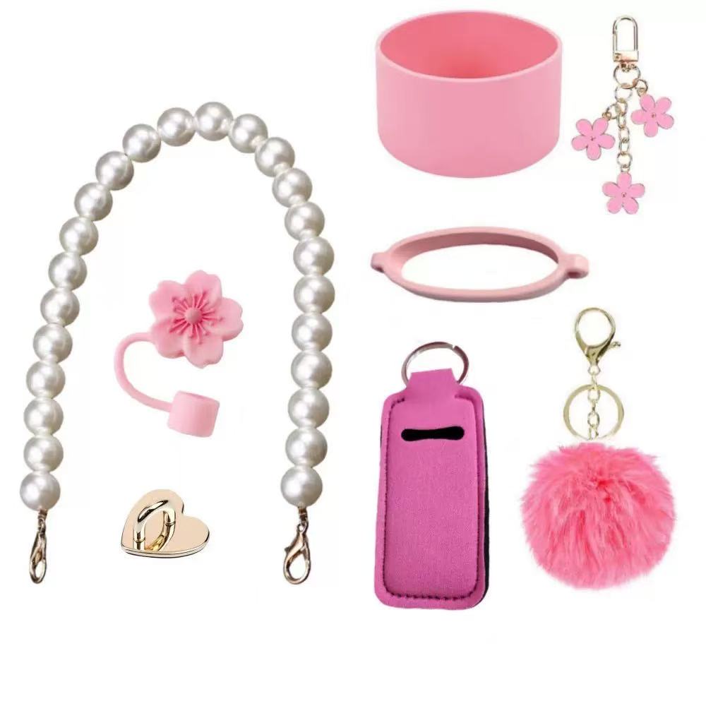 Pink tumbler accessory gift set featuring a pearl-style carrying strap, silicone cup boot, flower straw topper, decorative charm ring, fluffy pom-pom keychain, and matching pink tumbler accessories for customizing drinkware.
