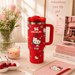 Red Stanley mug with Hello Kitty design on a table with flowers and books.