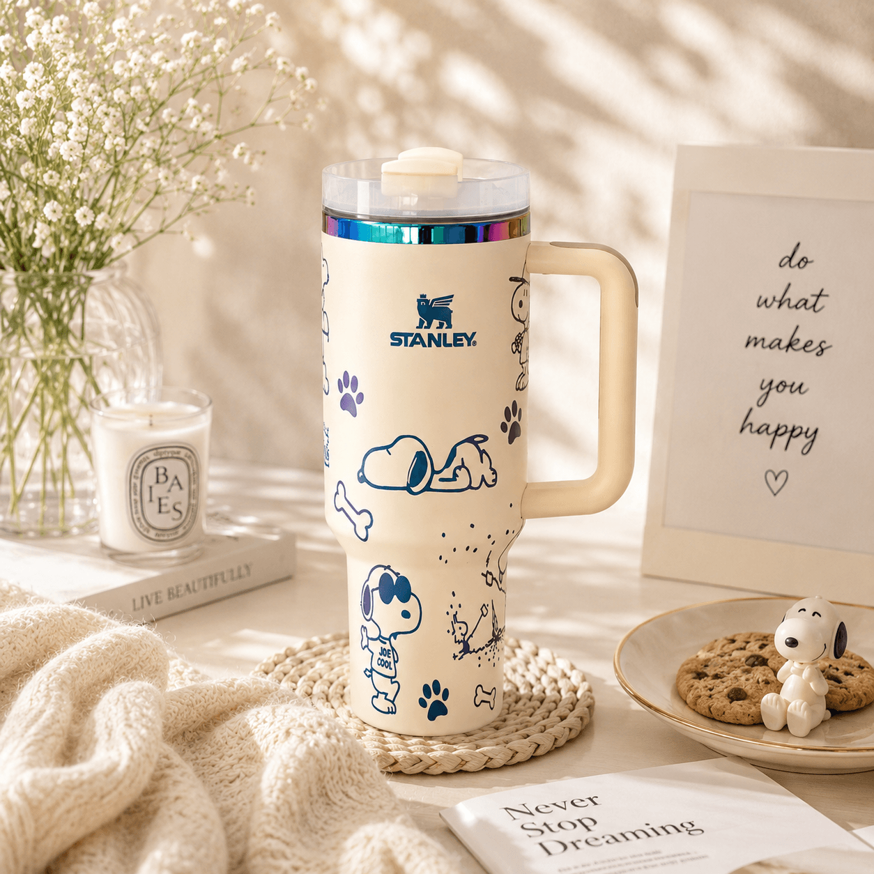 Stanley mug with Snoopy design on a table with a candle, book, and cookies.