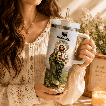 Person holding a Stanley mug with a religious design, surrounded by flowers and a framed quote.