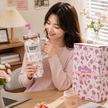 Woman holding a floral-patterned mug in a cozy room with a laptop, cake, and books.