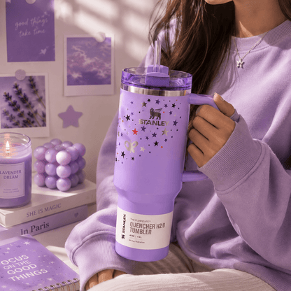 Person holding a purple Stanley tumbler with decorative elements in a cozy setting.